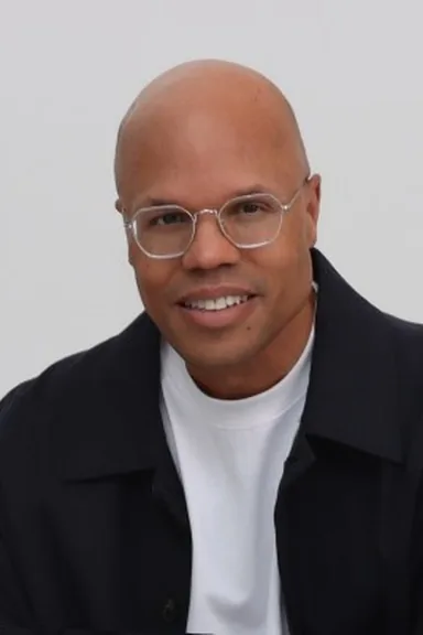 Hilmon Sorey in glasses, a dark jacket and a white T-shirt against a light background, smiling
