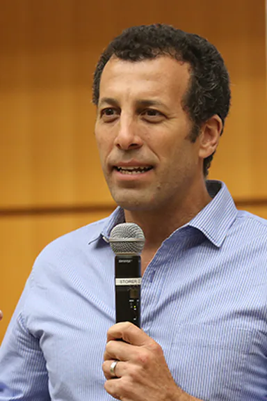 Jamie Rosenberg in a light blue shirt, microphone in hand, mid-sentence
