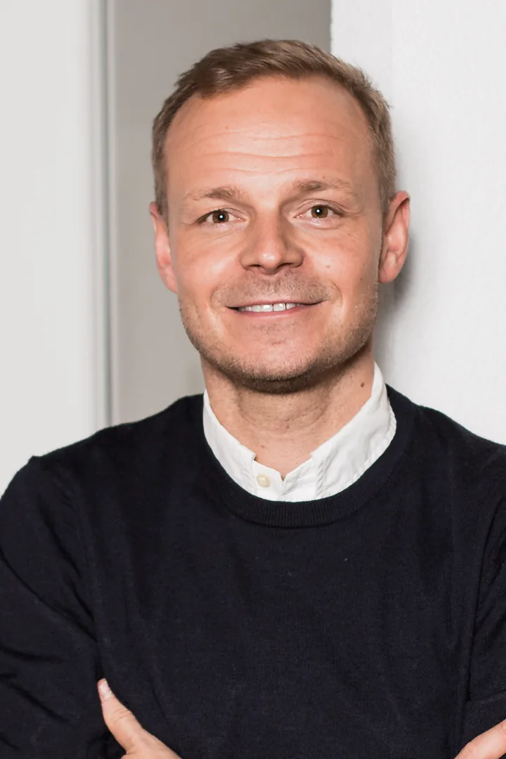 Konstantin Struck in a dark sweater over a white shirt, arms folded, leaning against a light wall